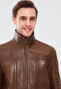 Giacca in pelle marrone con colletto rialzato, zip frontale e due tasche al petto con zip, caratterizzate da dettagli di cucitura orizzontale.