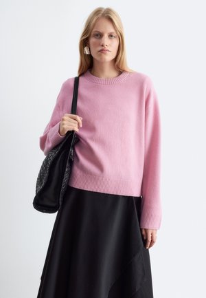 Jeune femme portant un pull rose et une jupe noire tient un sac à main noir tressé sur son épaule devant un fond uni.
