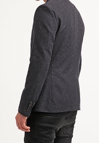 Granatowa marynarka ze strukturą, z wzorem w kropki, dopasowanym krojem i czterema guzikami na mankiecie, pokazana od tyłu.