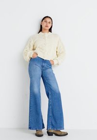 Cardigan a maglia cable cream con bottoni rotondi, abbinato a jeans a zampa d'elefante blu e stivali alla caviglia in pelle marrone con platform, il tutto contro uno sfondo bianco semplice.