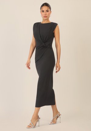 Schwarzes ärmelloses Kleid mit einem Twist-Detail an der Taille, figurbetonter Silhouette und knielanger Länge. Kombiniert mit silbernen High Heel Sandalen.