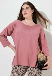 Rosa, lässig geschnittenes Oberteil mit rundem Ausschnitt und einer Brusttasche, kombiniert mit gemusterten braunen Hosen. Hält eine schwarze und naturfarbene gewobene Handtasche.