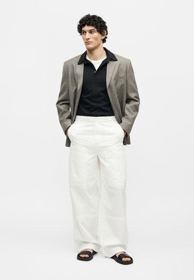 Νέος άνδρας στέκεται με τα χέρια στις τσέπες, φοράει γκρι σακάκι, μαύρο πουκάμισο, λευκό φαρδύ παντελόνι και μαύρα σανδάλια σε μονόχρωμο φόντο.