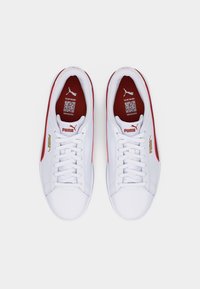 Ζευγάρι λευκά αθλητικά παπούτσια Puma με κόκκινες και χρυσές λεπτομέρειες, λευκά κορδόνια και ορατούς κωδικούς QR στις κόκκινες εσωτερικές σόλες, από πάνω.