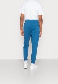 Blå joggare byxor i mjukt tyg, med ett elastiskt midjeband och ribbade muddar. En enda bakficka ger detalj.