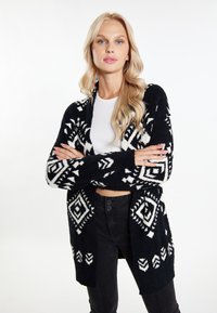 Pletený černobílý cardigan s geometrick�ými vzory, otevřený vpředu, s dlouhými rukávy a měkkou, texturovanou tkaninou. Kombinováno s bílým topem.