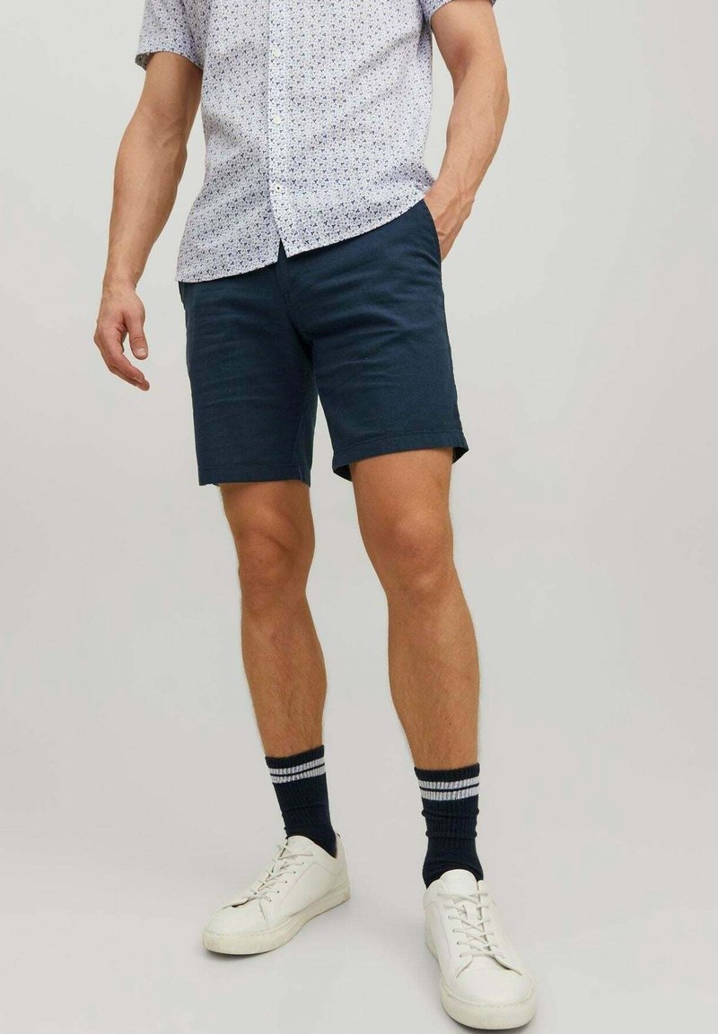 Homme portant un short bleu marine avec des chaussettes noires à rayures, des baskets blanches et une chemise à manches courtes à motifs boutonnée, debout avec une main dans la poche.