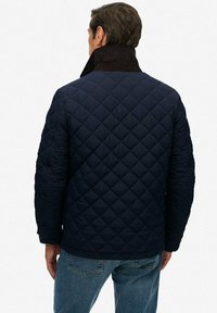 Chaqueta acolchada azul marino con cuello de pana, que presenta un exterior texturizado y un diseño sencillo. Combinada con jeans de un azul claro.