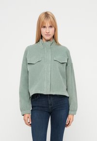 Veste en velours côtelé verte avec un col montant, deux poches poitrine et des poignets élastiqués, associée à un jean foncé sur un fond uni.