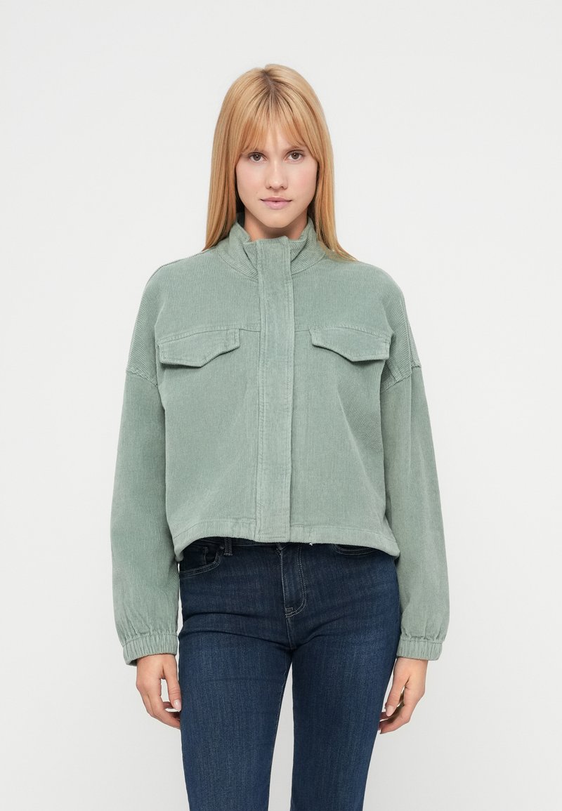 Veste en velours côtelé verte avec un col montant, deux poches poitrine et des poignets élastiqués, associée à un jean foncé sur un fond uni.