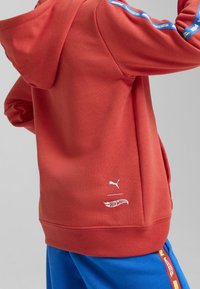 Roter Kapuzenpullover mit weißem Puma- und Hot Wheels-Logo auf dem Rücken, kombiniert mit blauen Hosen, die einen roten und gelben Seitenstreifen mit Puma Hot Wheels haben.