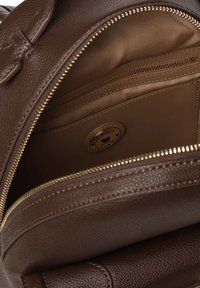 Interior de una mochila de cuero marrón con forro de tela color beige, que cuenta con un bolsillo con cremallera y una placa de logo redonda. Detalles de cremallera dorada visibles.