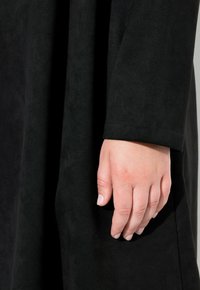 Schwarzer Wildlederstoff mit leichtem Glanz, der elegant fällt. Eine Hand liegt auf dem Material und zeigt eine glatte Textur sowie einen natürlichen Nagellack.