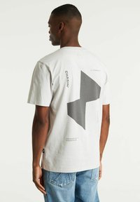 Lichtgrijze katoen t-shirt met een groot donkergrijs geometrisch ontwerp op de achterkant, met extra tekst en accenten op de stof gedrukt.