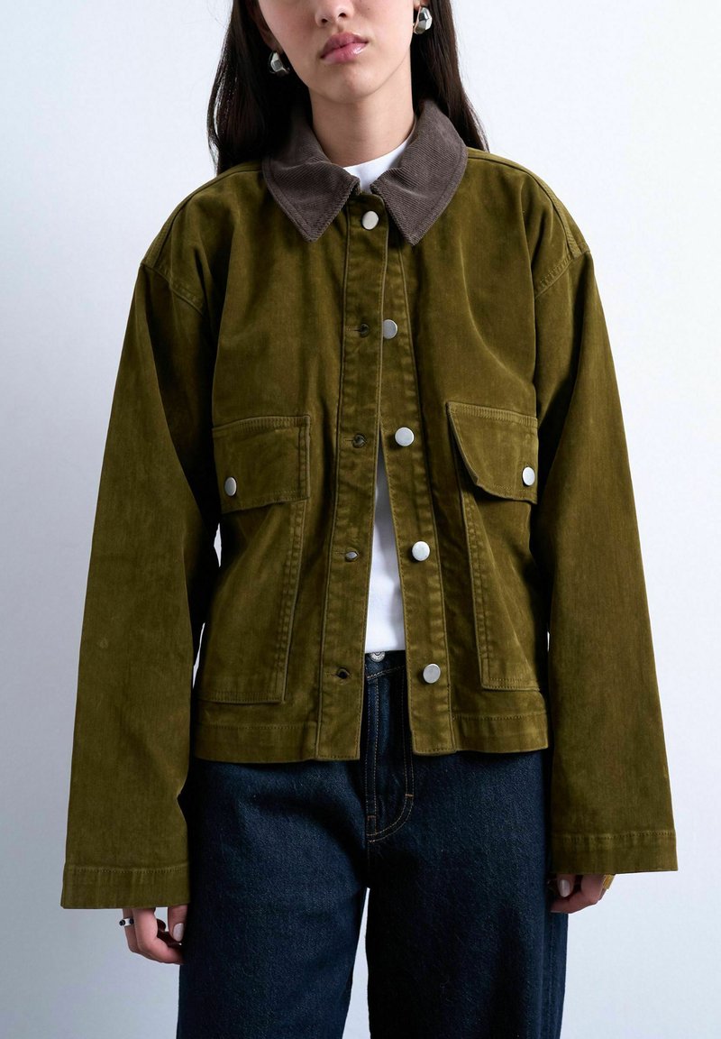 Femme portant une veste en velours côtelé vert olive avec col marron, boutons-pression argentés, poches à rabat à l'avant, chemise blanche et jean bleu foncé.