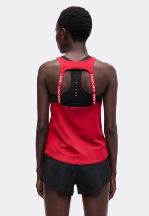 Atletická osoba nosiaca červené tielko Nike Pro cez čiernu športovú podprsenku a čierne šortky, zobrazená zozadu na jednoduchom pozadí.