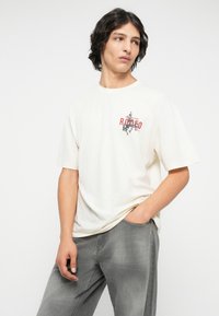 Kratki kremasti T-shirt s grafičkim dizajnom u crvenoj i crnoj boji na kojem piše "RODEO." Nositi s sivim hlačama, koje imaju opušteni kroj.