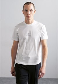 Camiseta de algodón blanca de mangas cortas con un sencillo dibujo en línea azul de manos formando un corazón en el pecho.