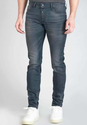 Jean slim fit gris foncé avec décoloration subtile, design à cinq poches, fermeture boutonnée et détails brodés. Porté avec des baskets blanches.