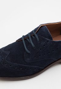 Navy modré semišové šněrovací boty s brogue designem, se sladěnými tkaničkami a hnědým koženým lemem na okraji kotníku.