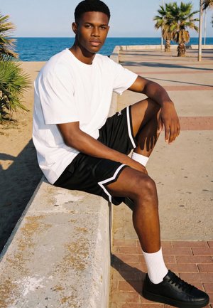 Młody mężczyzna w białej koszulce i czarnych szortach siedzi na betonowym murku w pobliżu palm i oceanu, patrząc w stronę kamery.