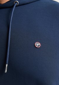 Hanorac bleumarin din material moale, cu un logo brodat în roșu și alb, având detalii cu un ochi metalic și șnur.