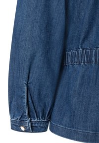 Dunkelblaue Denimjackenärmel mit gerafftem Detail am Bund, akzentuiert durch einen runden silbernen Knopf. Glatte Textur und schlichtes Design.