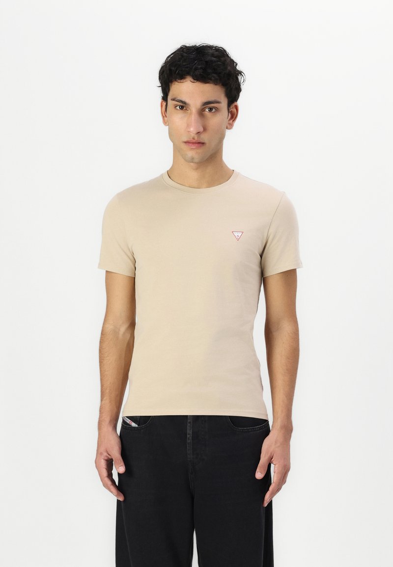 Camiseta de manga corta beis hecha de algodón suave. Presenta un pequeño logotipo triangular rojo en el pecho izquierdo. Se lleva con pantalones negros.