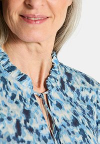Blau bedruckte Bluse mit gerafftem Ausschnitt und Bindeschluss. Sie verfügt über einen fließenden Stoff mit verschiedenen Blautönen und dezenten Mustern.