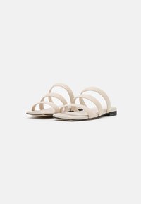 Sandali piatti beige con tre cinturini paralleli, una superficie liscia e un tacco nero basso. Il design è minimalista e con punta aperta.