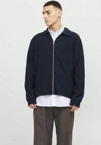 Navy blaue, gerippte Zip-Jacke mit Kragen, langen Ärmeln und geradem Saum, über einem weißen Hemd mit Knopfleiste getragen.