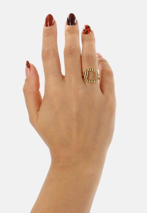 Anello d'oro con fascia intrecciata e design ovale aperto, indossato su una mano con unghie curate che presentano una combinazione di tonalità marroni e motivi tartarugati.