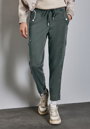 Personne portant un pantalon cargo vert avec une taille à cordon de serrage et des baskets beiges et blanches, debout contre un mur et un sol gris.