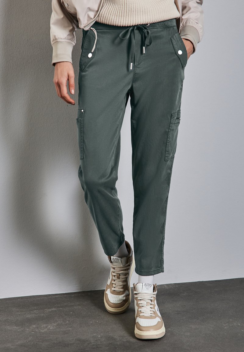 Cargo-byxor i mörkgrön bomull, med sidofickor med knappar, midja med elastisk dragsko och avsmalnande ben. Bärs med beige högsneakers.