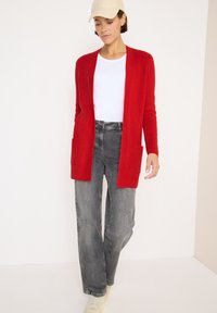 Rote gerippte Strickjacke mit offenem Vorderteil und zwei Taschen, getragen über einem weißen T-Shirt und grauen Jeans. Helle Sneaker runden das Outfit ab.