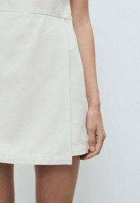 Cremfarbnes Kleid mit tailliertem Oberteil und schwingendem Rock. Sichtbare Nähte und eine glatte Textur. Hand ruht auf dem Rand des Rocks.