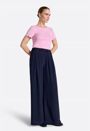 Donna in piedi con le mani dietro la schiena, che indossa un top rosa a maniche corte a coste e pantaloni blu navy a vita alta e gamba larga.