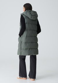 Frau trägt eine lange, olivgrüne, gepolsterte Weste mit Kapuze über einem schwarzen Langarmoberteil, dunklen Jeans und weißen Sneakers. Sie steht mit dem Rücken zur Kamera.