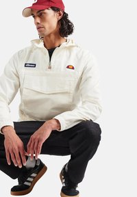 Biała kurtka z kapturem na zamek błyskawiczny, z przednią kieszenią i haftowanym logo. Czarna spodnie i czarne sneakersy z białymi paskami dopełniają zestaw.