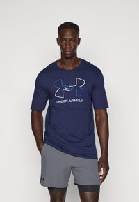 Navy blue short-sleeve T-shirt with a large white and blue Under Armour logo. Worn with gray shorts, showcasing a lightweight fabric.