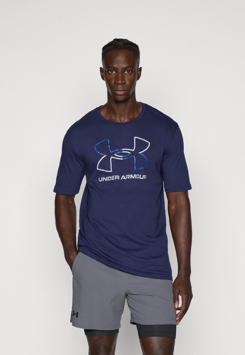 Mornarsko modra majica s kratkimi rokavi z velikim belim in modrim logotipom Under Armour. Oblečena z sivimi kratkimi hlačami, ki prikazujejo lahkotno tkanino.