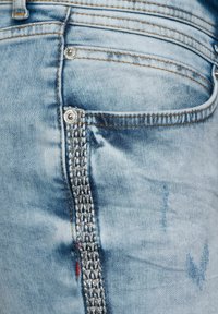 Světle modré džíny z denimu mají texturovaný vertikální akcent po straně, stříbrné kovové prvky a kontrastní stehy po celém oděvu.