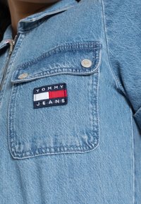 Ljusblå denimjacka med dragkedja, bröstficka och en rektangulär broderad Tommy Jeans-logotyp i rött, vitt och marinblått.
