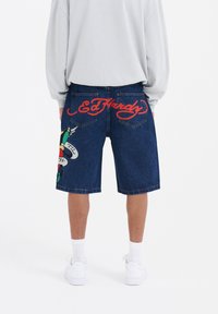 Pantalones cortos de mezclilla de corte holgado, color azul oscuro, con el logo rojo "Ed Hardy" y gráficos coloridos inspirados en tatuajes en el lado izquierdo.