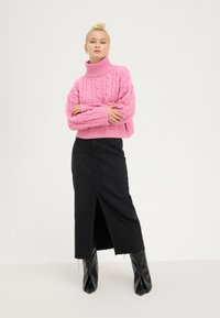 Pull en maille côtelée, cropped, couleur rose vif ; associé à une jupe en denim noir avec une fente à l'avant ; porté avec des bottes noires brillantes.
