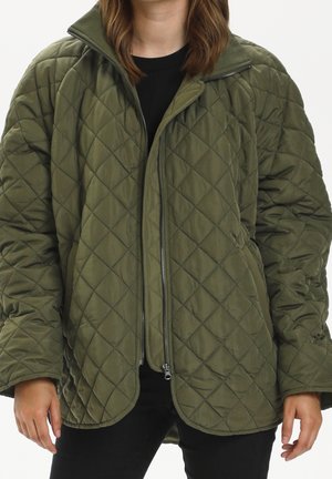 Nainen pukeutunut oliivinvihreään tikattuun takkiin, jossa on vetoketjulla suljettava etuosa, ja musta paita alla, seisomassa kädet rentoina sivuilla.