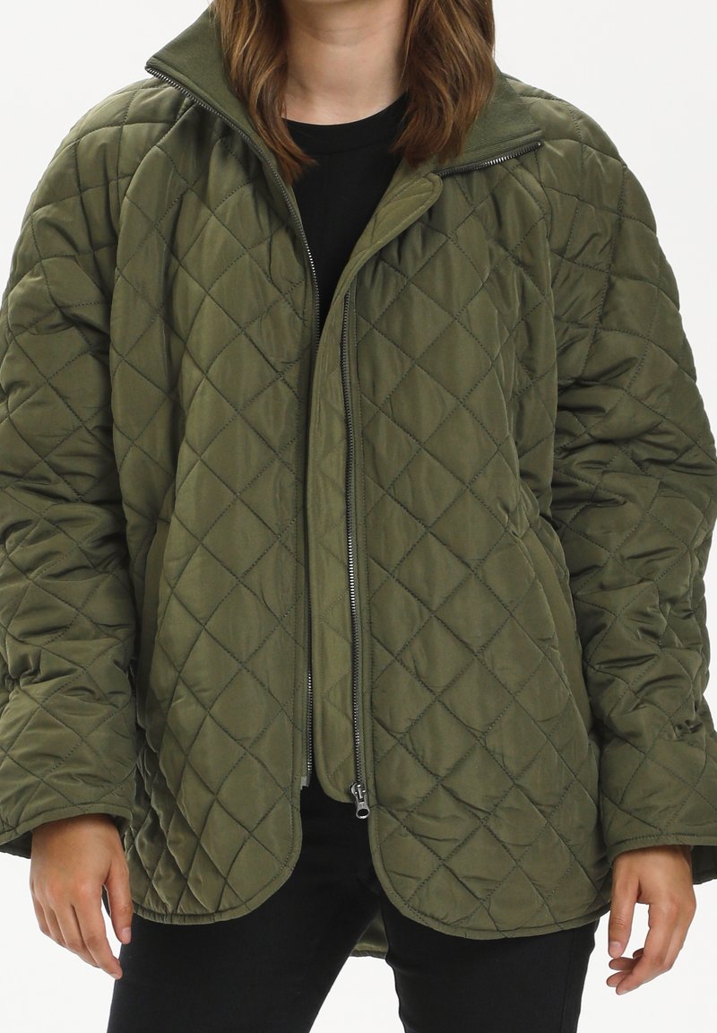 Femme portant une veste matelassée vert olive avec une fermeture éclair à l'avant et un chemisier noir en dessous, debout les mains détendues le long du corps.