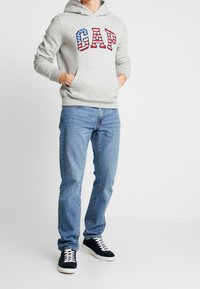 Sudadera gris con un gran logo "GAP" en rojo, blanco y azul, bolsillo tipo canguro, combinada con jeans azul claro y zapatillas oscuras.