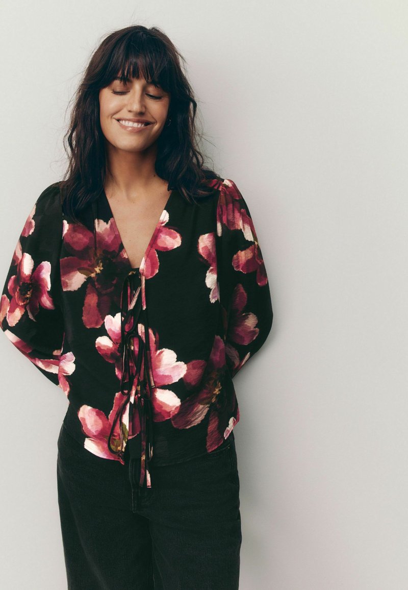 Blusa nera floreale con grandi fiori rosa e bianchi, scollatura a V, dettaglio frontale annodato e maniche larghe. Indossata con pantaloni neri.