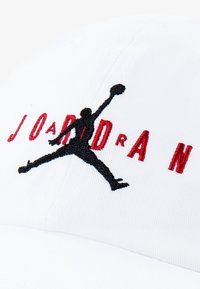 Vit basebollkeps med svart och röd broderad "JORDAN" och silhuettlogotyp. Har textilerad bomull och en böjd skärm.